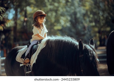 
Little Girl Riding a Horse Wearing Safety Helmet. Happy kid caring for her friend animal learning to ride it 
