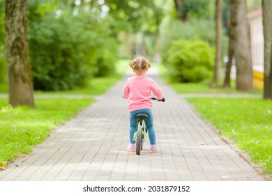 Little girl riding on first bike without pedals on sidewalk at city park in summer day. 2 years old toddler. Back view. Learning to keep balance.
