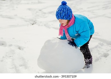 Little Girl Rolls A Big And Heavy Snowball To Build A Snowman In Winter Day