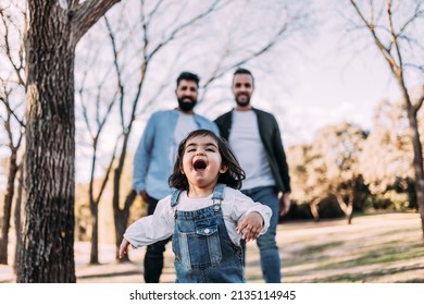 A Little Girl Running Happily Through The Park Followed By Her Parents. Gay Family Concept.