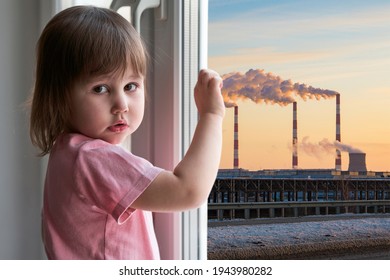 A little girl with a sad face stands at the window, outside the window is a bleak technogenic urban landscape with smoking factory chimneys. - Shutterstock ID 1943980282