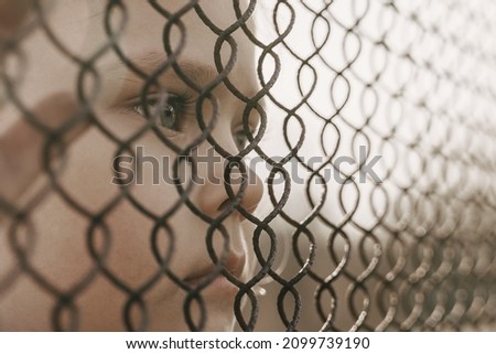 A little girl with a sad look behind a metal fence. Social problem of refugees and forced migrants.