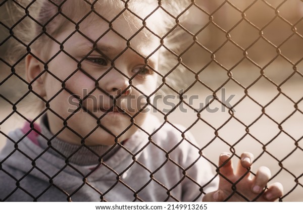 A little girl with a sad look behind a metal\
fence. The problem of forced deportation of Ukrainian children to\
Russian-occupied territories.