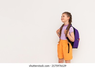 A Little Girl With A Satchel On Her Back Looks At Your Advertisement On A White Isolated Background. Summer Courses For Children. Copy Space.