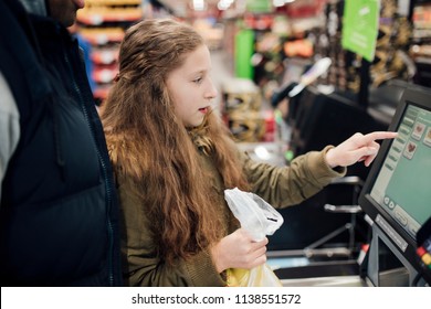 Little Girl Is At The Self Service Checkout Of The Supermarket With Her Father. 