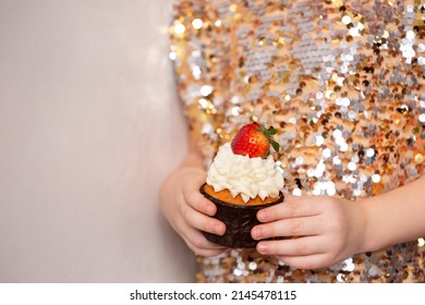 Little girl in a shiny dress with sequins, holding a handsome cupcake, close-up. Delicious cupcake on a sparkling background. Birthday party. Children's party. Selective focus on strawberry