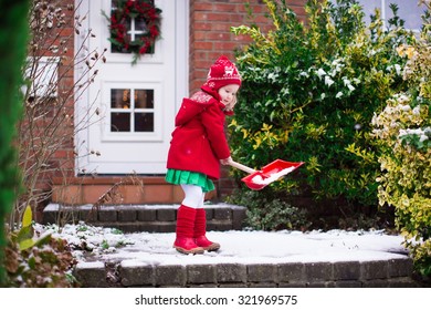 Little Girl Shoveling Snow On Home Drive Way. Beautiful House Decorated For Christmas. Child With Shovel Playing Outdoors In Xmas Season. Family Removing Snow During Blizzard. Kids Play Outside.