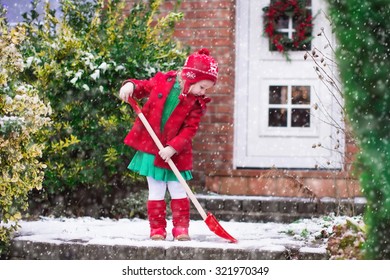 Little Girl Shoveling Snow On Home Drive Way. Beautiful House Decorated For Christmas. Child With Shovel Playing Outdoors In Xmas Season. Family Removing Snow During Blizzard. Kids Play Outside.