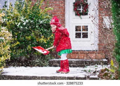 Little Girl Shoveling Snow On Home Drive Way. Beautiful House Decorated For Christmas. Child With Shovel Playing Outdoors In Xmas Season. Family Removing Snow During Blizzard. Kids Play Outside.