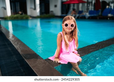 Little girl sits by poolside in pink dress, sunglasses. Smiles on sunny day, feet dipping in water. Kid enjoys luxury resort, summer vibes. Fun childhood moment, family vacation concept. - Powered by Shutterstock