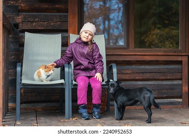 A little girl sits with a cat and a dog on the porch of a country wooden house.