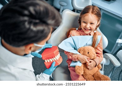 A little girl sits in a dental chair, holds a toy rabbit in her hands, laughs and looks at the dentist who shows her a model of the jaw with teeth. Children's dentistry. - Powered by Shutterstock