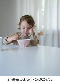 a little girl sits in the kitchen at a round table barefoot in a bright house in the kitchen and eats with an appetite organic cereals with vegetable milk, drinks clean water                          