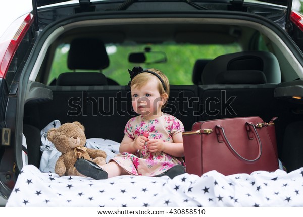 The little girl\
sits in luggage carrier of the family car. Near her there is bag\
and the bear Teddy sits.