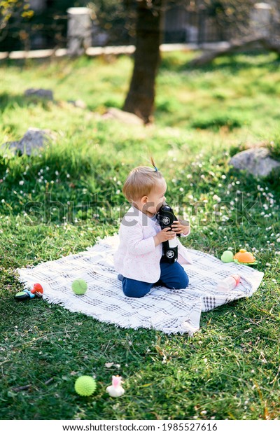 Little girl sits on a blanket on the green grass and\
chews a toy car. Side\
view