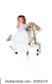 Little Girl Sits On A Carousel Horse Wearing A Long White Dress.  She Is Sucking Her Thumb And Dreaming In Her Own Little World.  Hairbow And Curls.