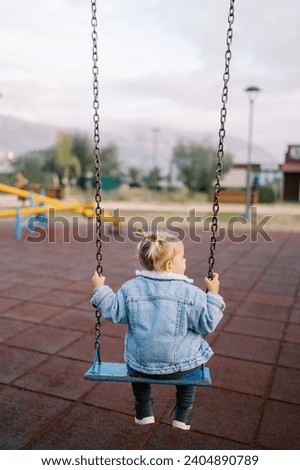 Little girl sits on a chain swing in the playground and looks away. Back view