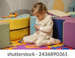 Little girl sits on colorful play mat, fully absorbed in solving handheld puzzle toy in preschool environment.