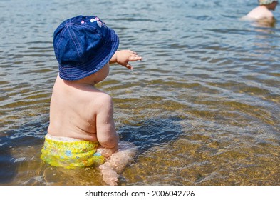 little girl sits on her bottom in yellow swimming trunks and a blue panama hat in the water in the river, pointing her finger into the distance