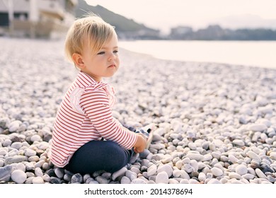 Little Girl Sits On A Pebble Beach And Looks At The Sea. Side View