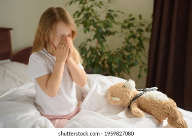 A little girl sits and sneezes on the bed among the blankets and pillows. House dust mite allergy - Shutterstock ID 1959827080