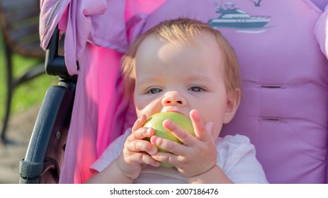 A Little Girl Sitting In Baby Carriage With A Big Appetite Eats An Apple.