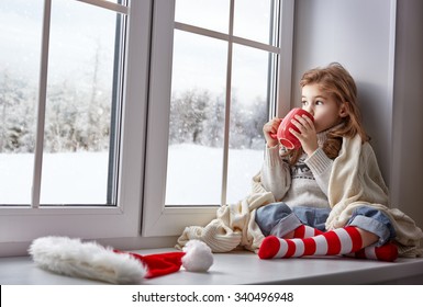 little girl sitting by the window with a cup of hot drink and looking at the winter forest
