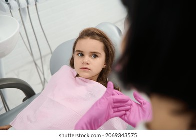 Little Girl Sitting In Dentist Chair, Listening A Paediatric Hygienist Teaching Her About Oral Hygiene And Prevention Of Baby Teeth Decay During Dental Visit In White Clean Modern Medical Cabinet