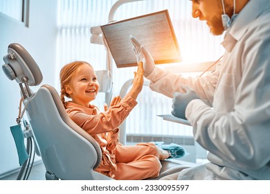 A little girl is sitting in a dentist's chair, giving a high five to the doctor and laughing. Dental care, trust and patient care. Children's dentistry.Sunlight. - Powered by Shutterstock