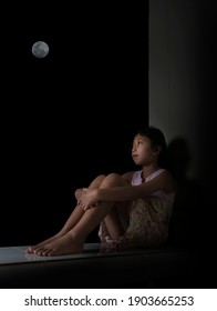 A Little Girl Sitting, Holding Her Knees, Looking Up At The Sky At Night, The Full Moon. Showing Feelings Of Loneliness And Sadness