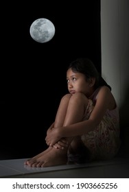 A Little Girl Sitting, Holding Her Knees, Looking Up At The Sky At Night, The Full Moon. Showing Feelings Of Loneliness And Sadness