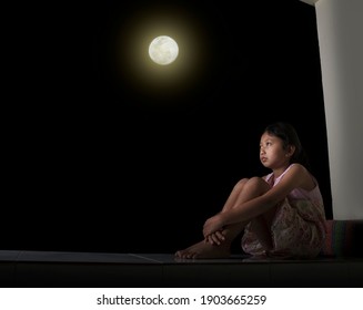 A Little Girl Sitting, Holding Her Knees, Looking Up At The Sky At Night, The Full Moon. Showing Feelings Of Loneliness And Sadness