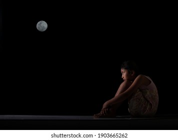 A Little Girl Sitting, Holding Her Knees, Looking Up At The Sky At Night, The Full Moon. Showing Feelings Of Loneliness And Sadness