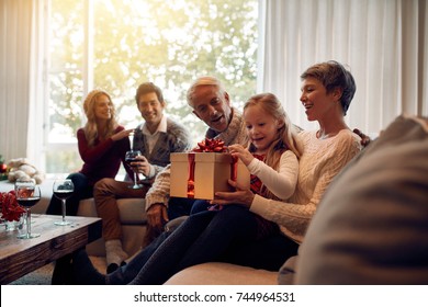 Little Girl Sitting In Living Room With Family Opening Christmas Gifts. Extended Family Gathering On Christmas Eve.