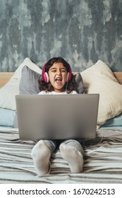 Little Girl Sitting On The Bed Between Cushions Laughing While Playing Or Watching A Movie On A Computer, Has A Pink Headset, Child And Technology Concept, Copy Space For Text