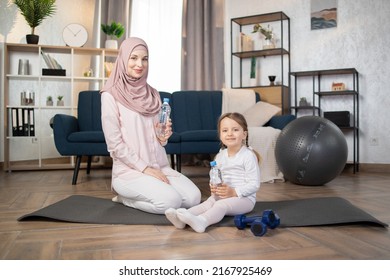 Little Girl Sitting On Mat With Her Young Muslim Mom While Doing Training Morning Exercises At Home. Small Kid Doing Sports Healthy Practice Of Yoga, Pilates With Mother Holding Bottles With Water.