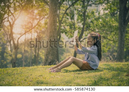 Little girl sitting on meadow  using computer tablet or smartphone in park, vintage tone photo