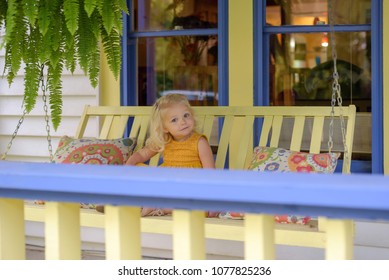 Little Girl Sitting On A Porch Swing On The Front Porch On A Summer Day