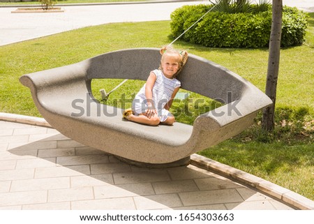 A little girl is sitting on a stone bench in a city Park and has her hands over her eyes against the bright summer sun.
