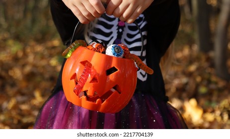 Little Girl In Skeleton Costume Holds In Hands Orange Pumpkin Plastic Bucket Full Of Assorted Candies And Jelly Worms. Halloween Trick Or Treat Concept. Banner, No Face, Unrecognizable. Forest Behind.