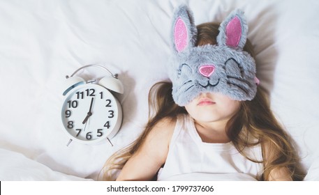 Little girl sleeping in bed under white blanket wearing grey bunny plush sleep mask with alarm clock on pillow. Early morning wake up. Putting kid to sleep.Mom's correct daily routine, rest for child.