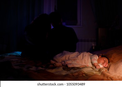 The Little Girl Sleeping On Bed Near The Hanging Parents Blurred On Back Side. Happy Family Concept. Night Time Dark Room. Selective Focus