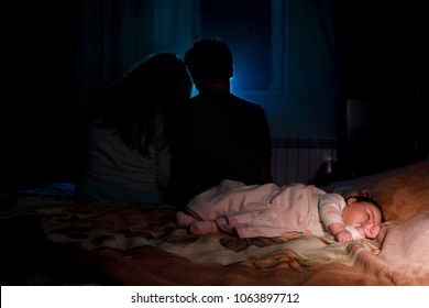 The Little Girl Sleeping On Bed Near The Hanging Parents Blurred On Back Side. Happy Family Concept. Night Time Dark Room. Selective Focus