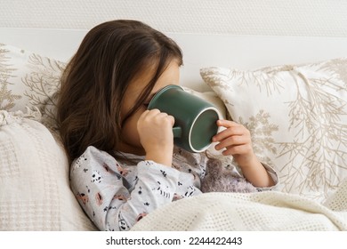 Little girl in sleepwear lie under blanket and take sip of drink covering face with mug. Portrait of unhealthy child, side view. Kid have bed rest. Home treatment, cold, disease. Taking medicament. - Powered by Shutterstock