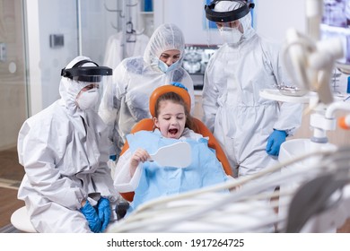 Little Girl Smiling Looking In Mirror Sitting On Dental Chair After Oral Hygine Procedure Dressed In Ppe Suit. Child Wearing Ppe Suit During Teeth Intervention At Dental Hospital.