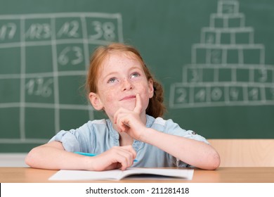 Little Girl Smiling As She Sits At Her Desk In Class Writing Notes Staring Up Into The Air With A Pensive Expression With The Blackboard Behind Her