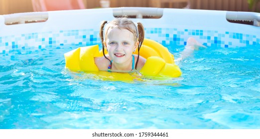 Little Girl Is Splashing In Swimming Pool Outdoor, Having Fun. Yellow Swimming Aids On Child. Above Ground Metal Frame Swimming Pool