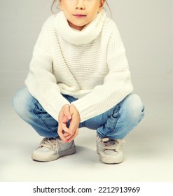 Little Girl Squatting, Wearing Jeans And Warm Sweater