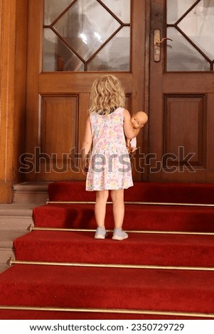 a little girl is standing with her doll on a staircase covered with a red carpet and leading up to a large door.