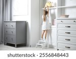 Little girl standing on step stool and reaching for toys on shelf indoors, back view